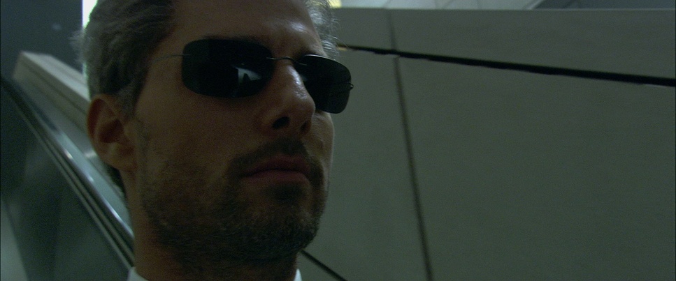  Close up of Tom Cruise riding down an escalator. He's stoic and wearing sunglasses.