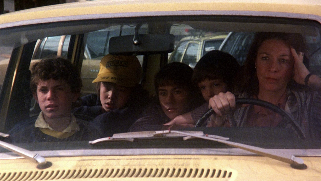 The interior of a yellow car, shot through its front windshield. Jane Alexander sits in the right of the frame, with her head resting against her hand and the other hand gripping  the car’s black steering wheel. She has brown shoulder length hair, and is wearing a black wristwatch, and what looks like a light colored poncho or a sweater. To her right sit four children: a little boy with brown hair whose face is slightly obscured by her steering wheel hand, a girl with brown hair and bangs, another boy in a yellow baseball cap, and a boy with brown curly hair and a yellow collar peeking out of a darker colored sweater. Jane looks worried and tense, and the children sit looking ahead. Behind them through the back windows you see pieces of two other late 1970s/ early 1980s era cars.