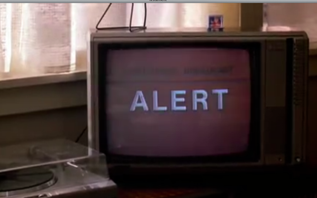 An old  television set’s screen is black, with the word “ALERT” going across it in big white capital letters. To the left and running behind it is the bottom of a white window sill with white curtains with daylight behind them. Below this, to the slide and slightly in front of the corner of the TV is a record player, the other half of which is cut off by the frame.