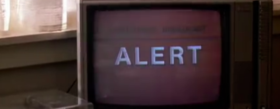 An old television set’s screen is black, with the word “ALERT” going across it in big white capital letters. To the left and running behind it is the bottom of a white window sill with white curtains with daylight behind them. Below this, to the slide and slightly in front of the corner of the TV is a record player, the other half of which is cut off by the frame.