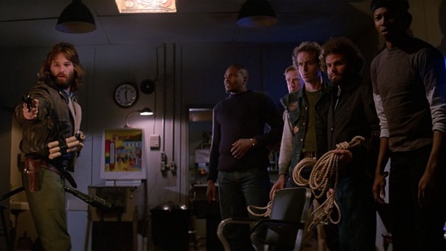 Kurt Russell as MacReady points a gun at something out of frame while the rest of the gang of Outpost 31 looks onward ready to respond.
