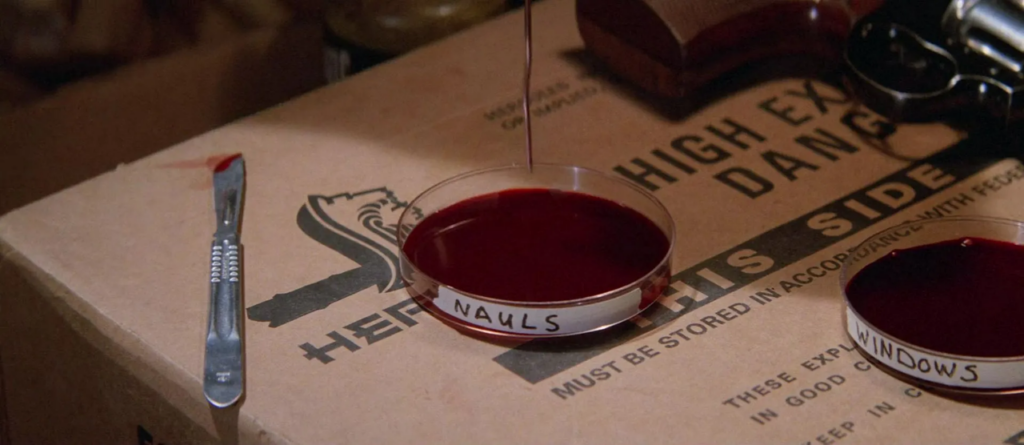 A petri dish filled with blood labeled “Nauls” sits in the center of a cardboard box, with another petri dish filled with blood labeled “Windows” sits to its right. To the left of both dishes is a blood scalpel.