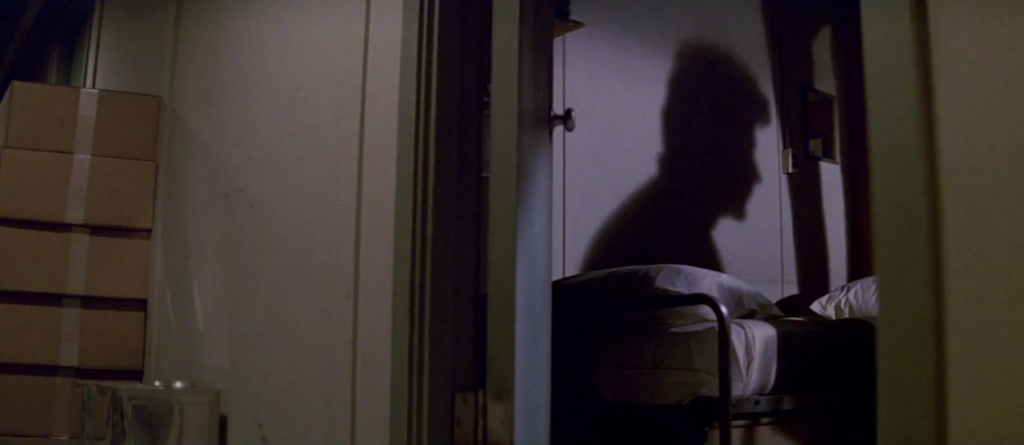 A view from the hallway of a mysterious figure’s shadow on the wall above a researcher’s bed.