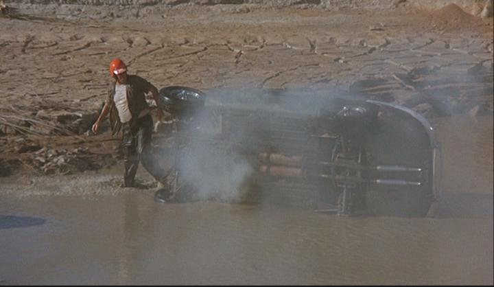 A man wearing a red helmet stands to the left of a crashed Jaguar car, which is tilted over in a bed of water, smoking.