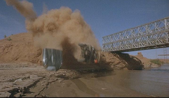 In the center of the frame, a silver car is falling off a cliff, the front cover detached. there is a massive cloud of dust covering the top of the image. to the right is a bridge, and at the bottom is a bed of muddy water.