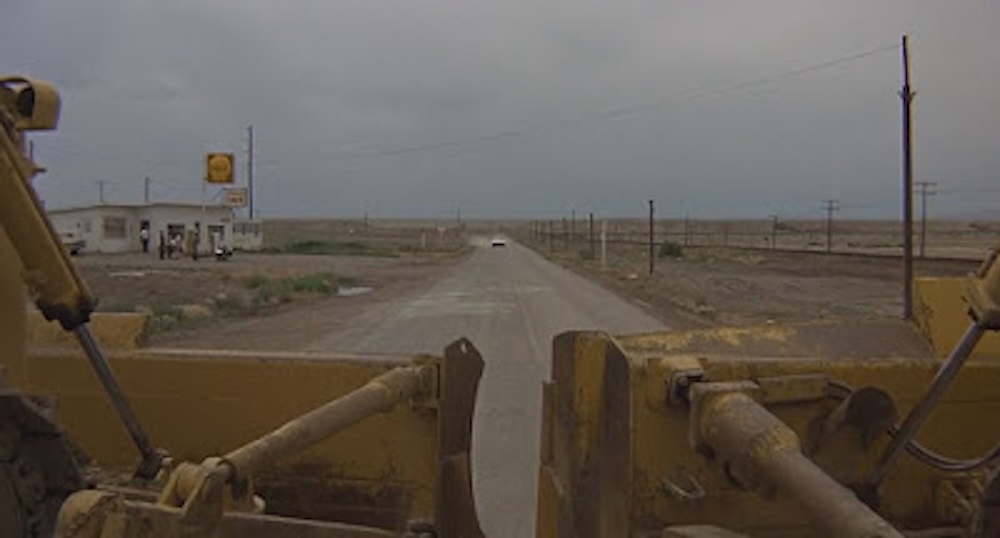 Two bulldozers face the road in the foreground of the image; in the distance is a car driving toward the bulldozers. to the left is a gas station with several people standing outside.
