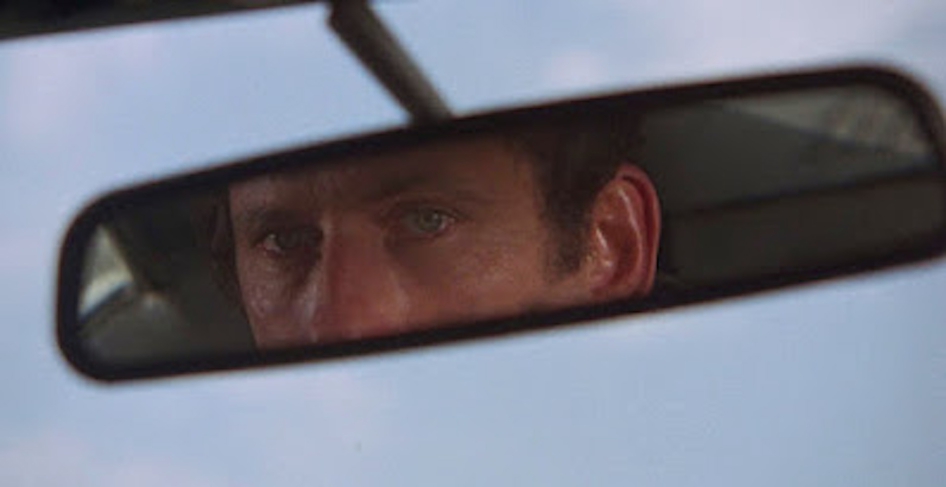 A close-up shot of a white man with green eyes looking at himself in the rearview mirror of his car.