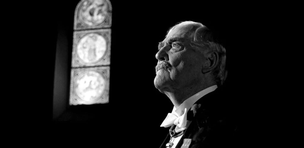 A semi-close up shot of a man in a tuxedo in a dark room; there is a lit up sign of three icons to his left. they are hard to distinguish but look like saints.