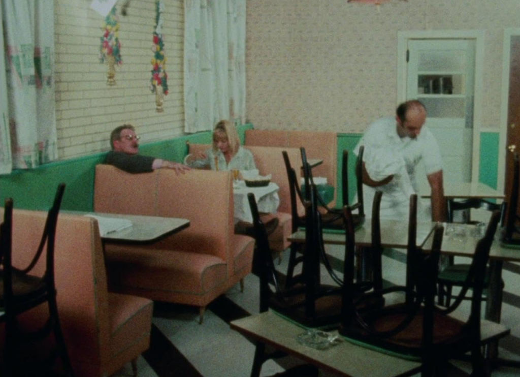 Wanda and Mr. Dennis sit in a pink booth at a restaurant while a server stacks chairs on the tables.
