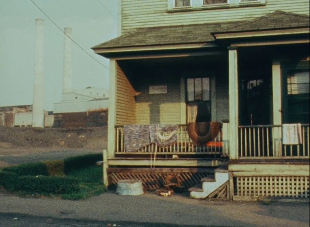 A long shot of a semi-dilapidated two-story home that fills the frame on the right yet reveals a factory with smoke stacks not too far off from the home on the left side. A row of sad, short shrubs line the one small green area of the image, slightly dividing the factory and the home and a powerline droops down from the upper left corner connecting to the house. A metal washtub sits outside the rundown wraparound porch, and blankets and rugs are left out to dry over the ledge above it. Imperfect latticework and a meager three-step stairway up to the porch help keep the home from inevitable floodplain dangers. Shadows overhang the gray shingles and windows, suggesting that either no one is home or no one is awake.