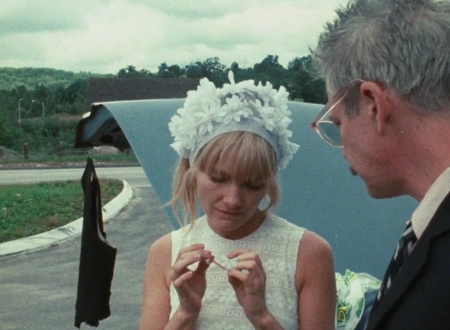 A medium close-up two show of Loden as Wanda in the center of the frame with Michael Higgins as Norman Dennis to her right, faced away from the camera and encroaching on her space, looking down at her nails that she is filing as they stand in front of Dennis's open gray-blue sedan's trunk. A nondescript item of clothing hangs from a wire off the suspended hatch, and the background suggests they have pulled off the side of a highway somewhere in middle America, a lush forest in the distance. Dennis is wearing a proper suit and tie while Wanda is slumped over, her head crooked as she files away, adorned in a white churchgoing sleeveless dress and white floral headpiece. Her scraggly blond hair peeks out around her face, as if screaming for help but also resigned to never find any.
