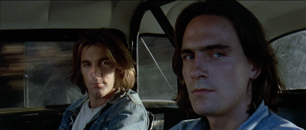 Two men sit in the front of a grey car. They are looking to frame right in the front seat. On the left is The Mechanic, played by Dennis Wilson. On the right is The Driver, played by James Taylor.
