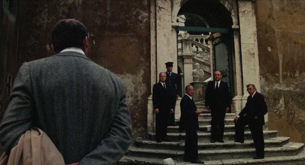 A man in a grey suit (Rosi), seen from behind, watches a group of five men in dark suits and one policeman gathered on stone steps at a large arched entrance.