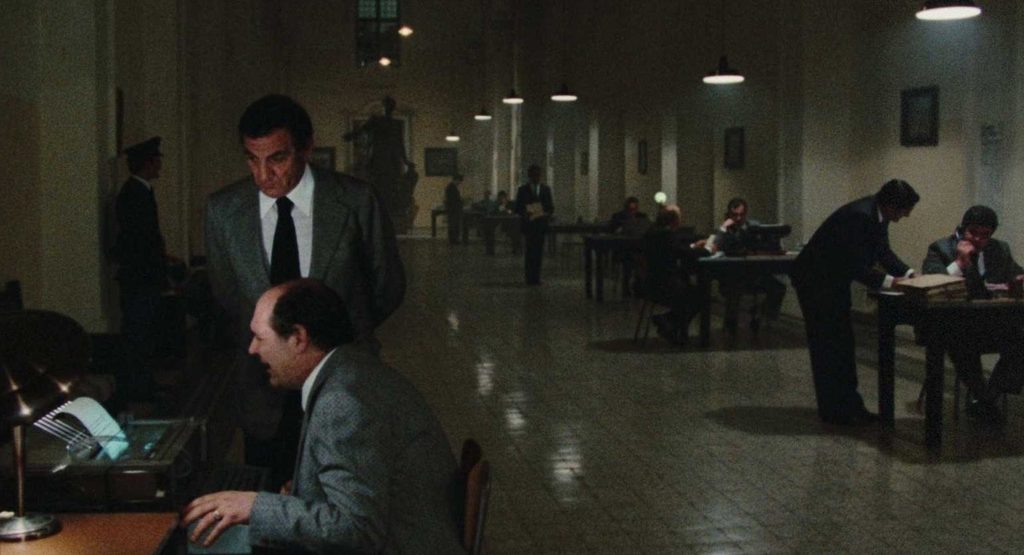 A medium shot of a large, dimly lit office at night, where a man in a tweed suit stands over another man typing at a desk, while other employees work further down the hall.