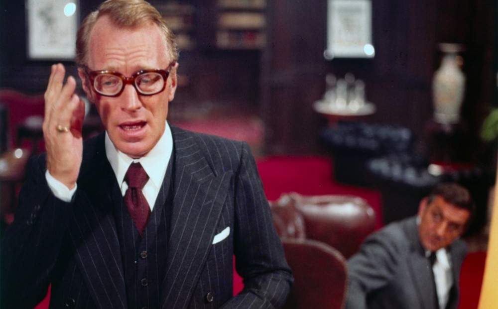 Max von Sydow, wearing a pinstripe three-piece suit and thick-rimmed glasses, speaks animatedly with his hand raised while Lino Ventura watches him from the background in a richly furnished office.