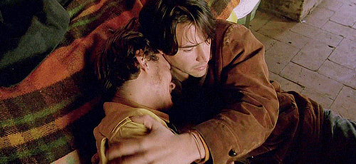 Two young men lean against the side of a bed. One cries into the other’s shoulder, who touches the man’s neck.