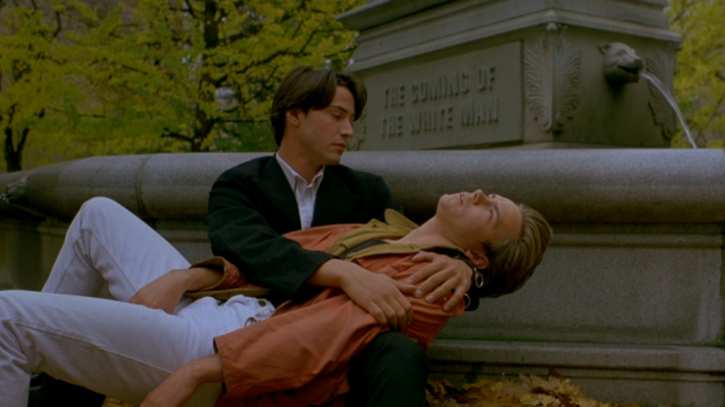 A young man sits against a statue in the park, inscribed with “THE COMING OF THE WHITE MAN.” He holds an unconscious blonde man across his lap.