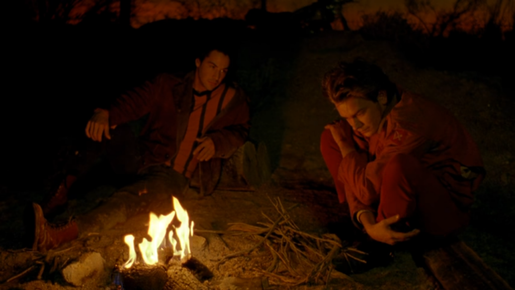 A young man hunched into a squat stares into a bonfire, wrapping his knees in his arms. Another man stares at him, sitting near with his leg extended. 
