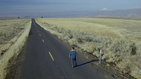  A man in a denim jacket stands in the middle of an empty desert highway, surrounded by fields and mountains.