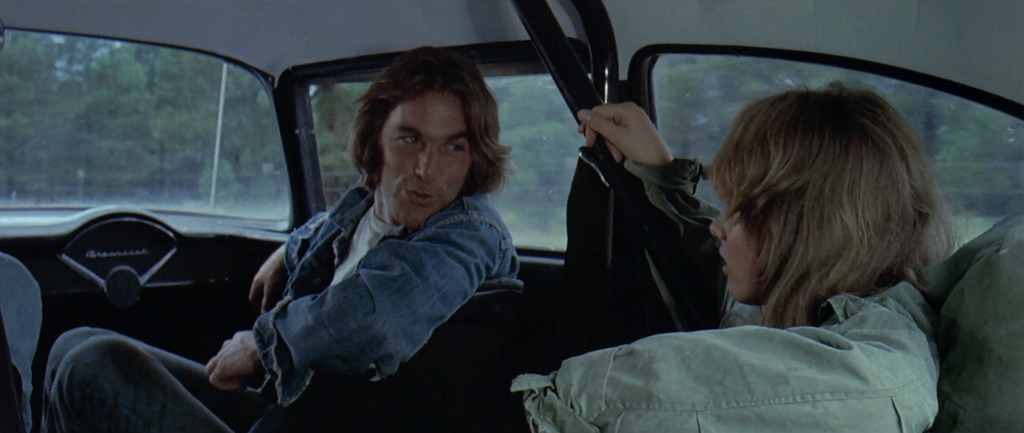 The Mechanic sits in the passenger seat. He is turned looking to the back seat, talking to The Girl, played by Laurie Bird, who is in the back seat in a denim shirt.