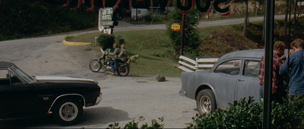 A black car sits on the left side of frame at a motel parking lot. To the right is the grey Chevy that the Driver and the Mechanic drive throughout the movie. A man in a red checkered shirt and a man in a blue shirt are looking at it. In the background a couple are sitting on a motorcycle and driving away. Behind them in the parking lot on the ground sits a duffel bag.
