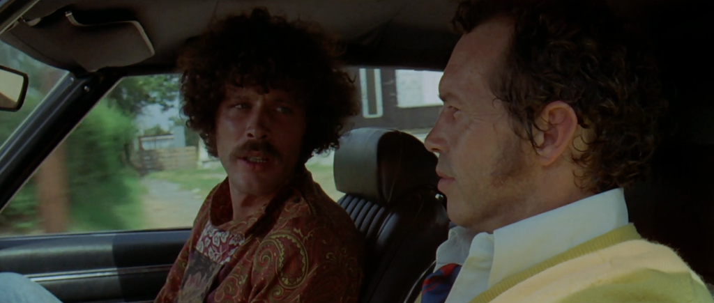 A man in a red paisley shirt and big curly hair and a mustache sits on the left side of the frame, sitting in the front seat of GTO’s car. He is turning and looking at GTO, a man in his 40s who is driving the car on the right side of the frame. GTO has an intense look on his face. He’s wearing a yellow sweater.