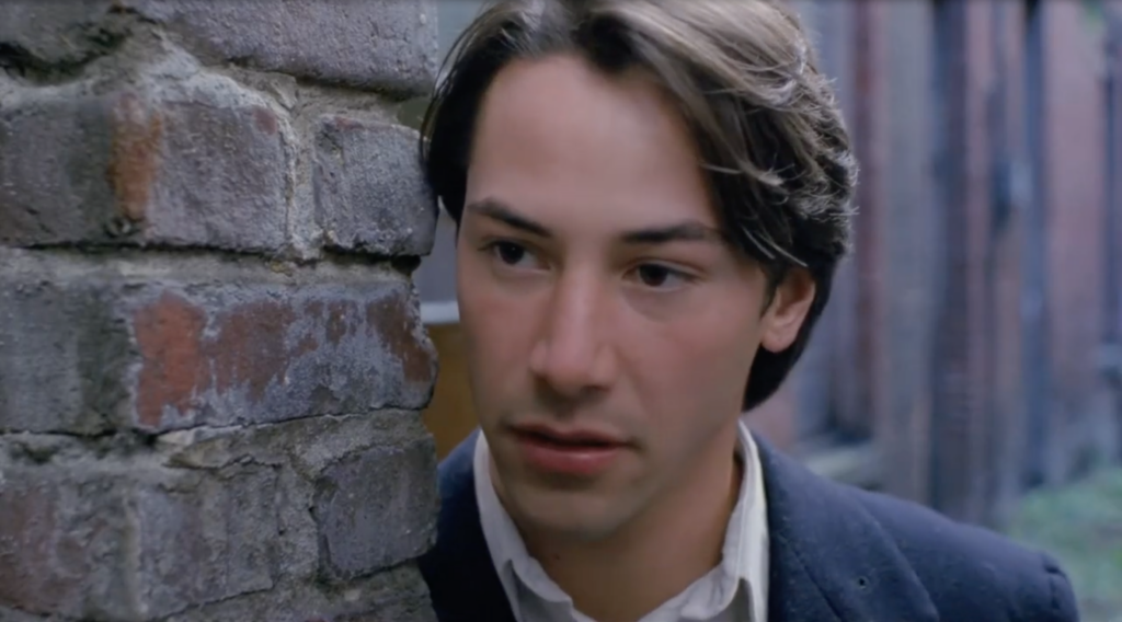 Keanu Reeves, dressed in a suit, leans his head against a brick door frame in Gus Van Sant's My Own Private Idaho.