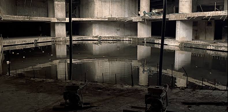 A large vacant building structure with a pool of water in the middle of the floor. From the film I Don't Want To Sleep Alone.