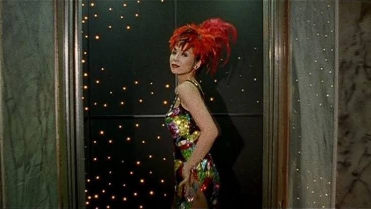 A Woman stands in a dingy elevator, but is dressed in exotic fashion as if in a Hollywood musical number.