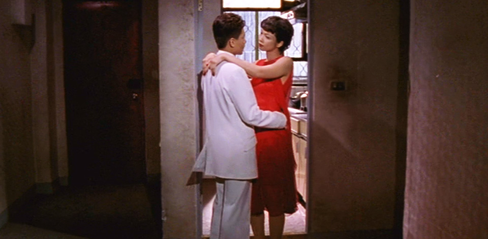 A Man in a white suit and a woman in a red dress dance together in the hallway of a dingy apartment.