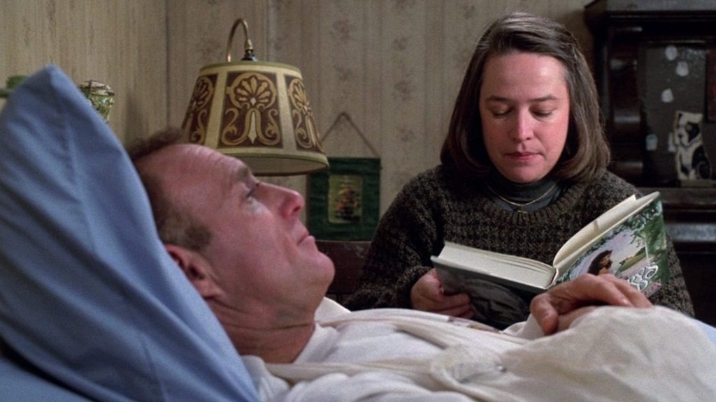 a white, brown-haired woman reading a book while sitting next to a white, blond man lying in bed