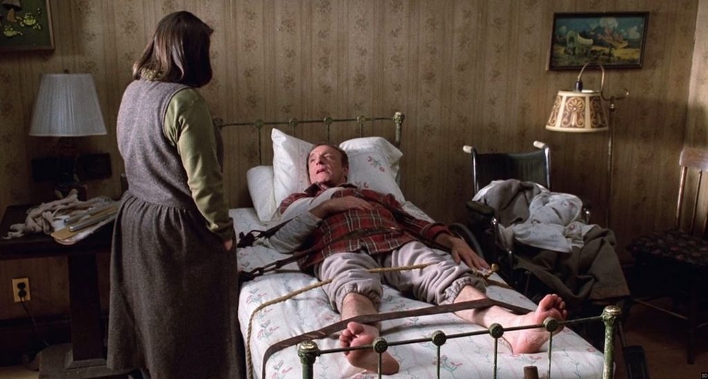 a white, brown-haired woman standing over a white, blond man tied to a bed