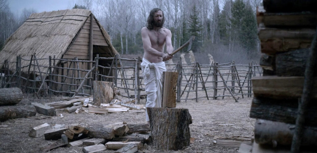 A shirtless white man with long brown hair stands in the center of a farm pen holding an axe above a piece of wood.