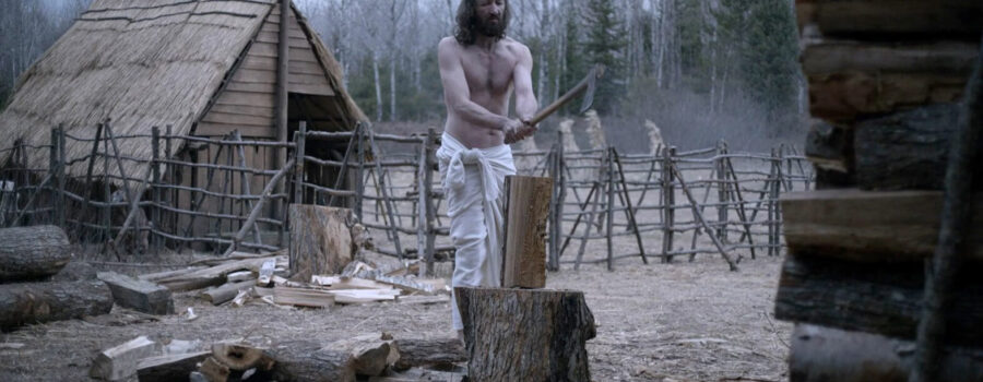 A shirtless white man with long brown hair stands in the center of a farm pen holding an axe above a piece of wood.