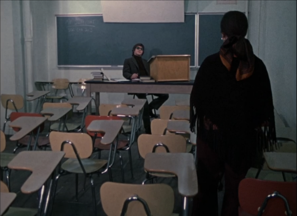 A man in sunglasses sits lazily by a podium at the front of a classroom, while a woman stands in a row of desks.