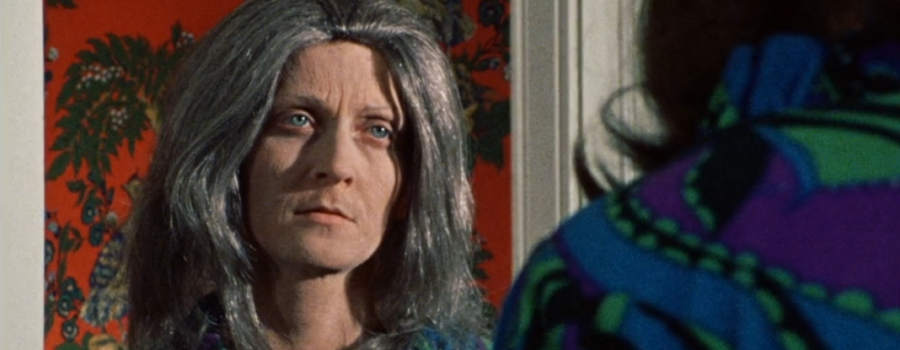 The female protagonist wearing a green and blue dress from the 1970s, meets eye to eye with an expressionless and elderly version of herself.