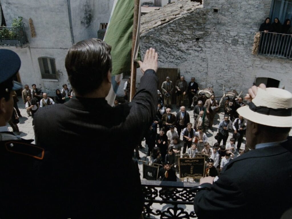 a white man with dark brown hair standing on a balcony saluting with one arm forward while a crowd of people looks up at him.