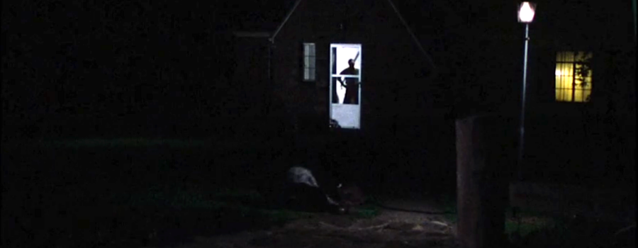 A woman lays on the ground in the front of a house. A man stands with his hands on his hips in the doorframe.