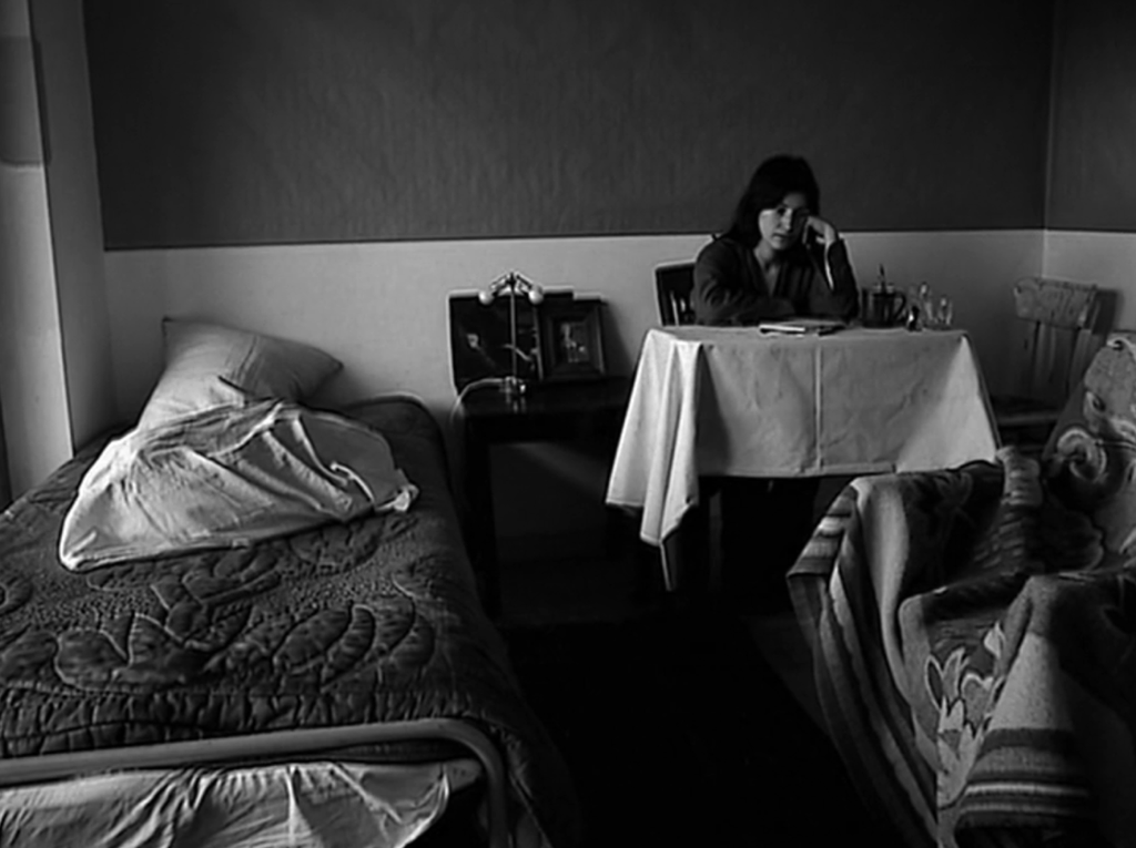Julie, a young brunette played by Chantal Akerman, sits slightly hunched over at a table off-center and in the background of the black-and-white frame. In the foreground of this small one-bedroom apartment are both her bed to the left and an armchair to the right, both of which are covered haphazardly with blankets. Two wrinkled pillows are strewn on the bed as well, and a black side table to its right, situated between the bed and the slightly largely table at which Julie is sitting. A white tablecloth hangs off Julie's table and some objects are laid on it. An armless wooden chair is also shoved into the corner to Julie's right. Julie's face is aimed slightly downward and resting on her fist, which is propped up on the table using her elbow. Her expression is one of blank but intense depression.