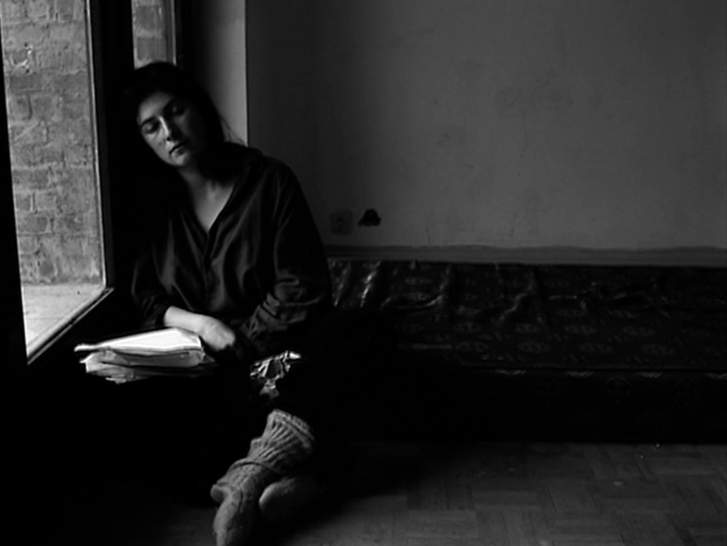 Julie is cross-legged on the corner of a mattress that lies in the corner on the floor of her apartment. She's leaning her shoulder and head on the floor-to-ceiling window frame on the left. Water damage to the wall in the background is clearly visible. Julie has a stack of disheveled papers on her right thigh, her hands resting in her lap, and her feet covered in thick wool socks in the foreground. A crinkled bag is also situated in her lap. She is once again looking slightly downward but with eyes closer to closed, suggesting a sleepy apathy or aimlessness. The mattress pad is rumpled and the patterned sheet is obviously worn. The wood floors are sprinkled with crumbs, visible from the light coming in through the window