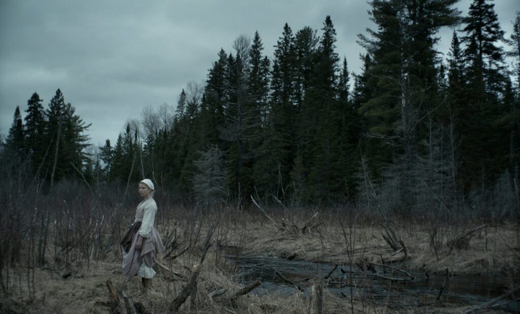 Wide shot of a woman in Puritan garb standing against a dark forest from Robert Eggers’s The VVitch.
