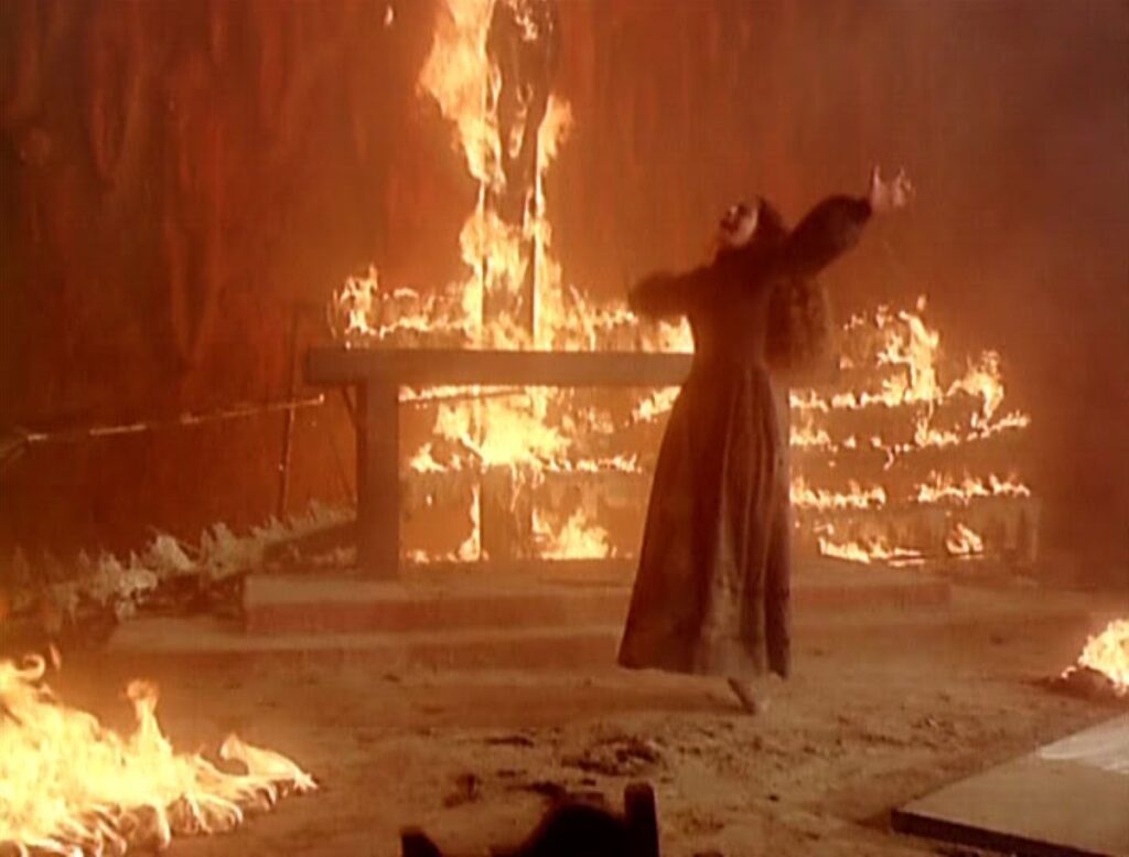 Wide shot of Alucarda (Tina Romero) in a long gown, throwing her arms open wide toward a blazing altar consumed by fire during the monastery inferno.