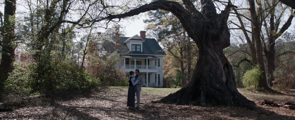 A man and a woman embrace outside of an older looking two story white house. They are under a curved, ominous looking tree