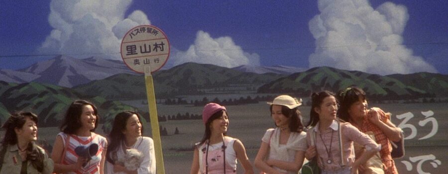 Seven young women named Kung Fu, Melody, Gorgeous, Fantasy, Sweet, Prof and Mac stand in front of a bus stop with a mountain landscape behind them with blue skies and some clouds.