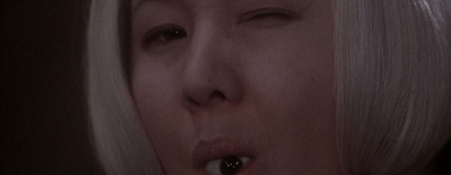 A closeup shot of a gray haired older woman winking at the camera with a detached eyeball in her mouth.
