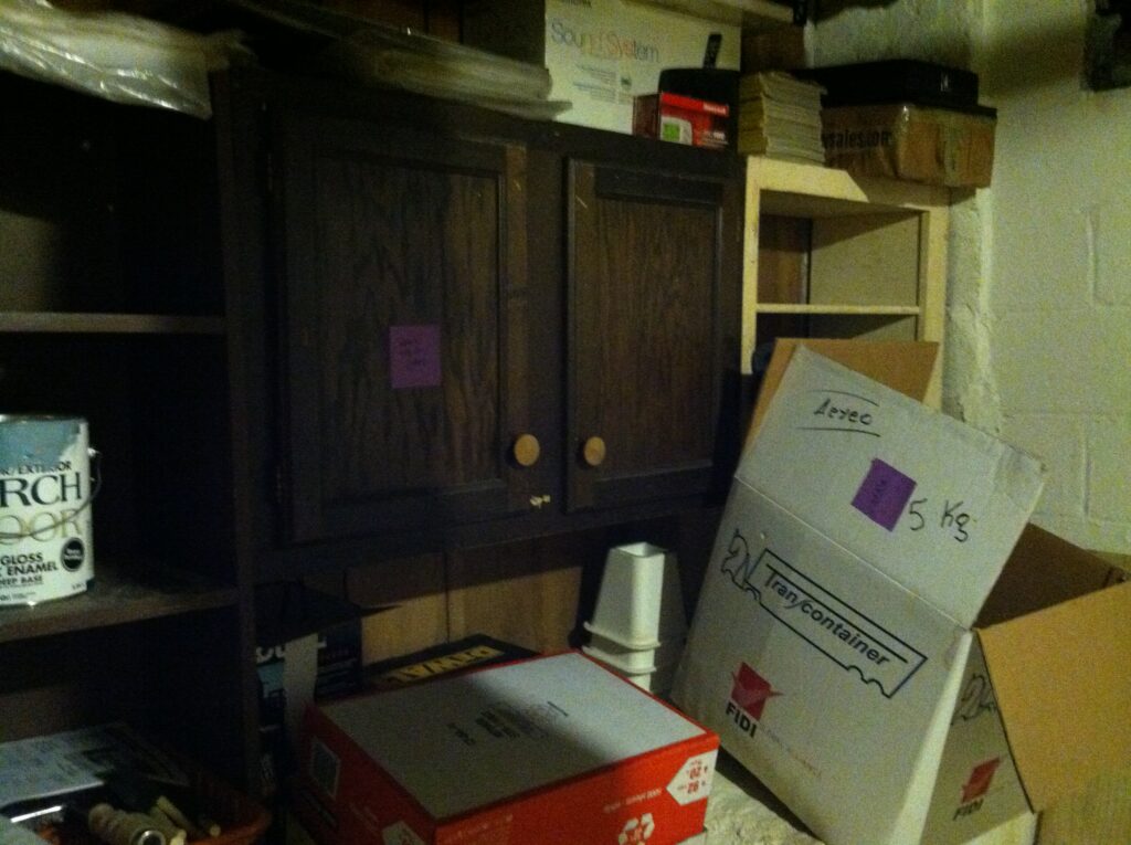 A brown cabinet on a wall, surrounded by cluttered stuff you would find in a basement. Boxes, cans of paint, etc. On it is a purple Post-It note.