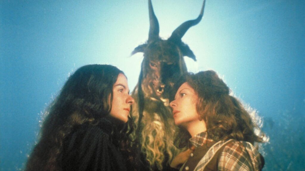 Justine and Alucarda gaze at each other in profile against a bright, hazy sky, standing before a towering figure in an eerie horned goat mask and pelt during a forest ritual.