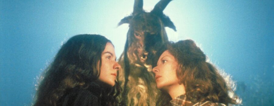 Justine and Alucarda gaze at each other in profile against a bright, hazy sky, standing before a towering figure in an eerie horned goat mask and pelt during a forest ritual.