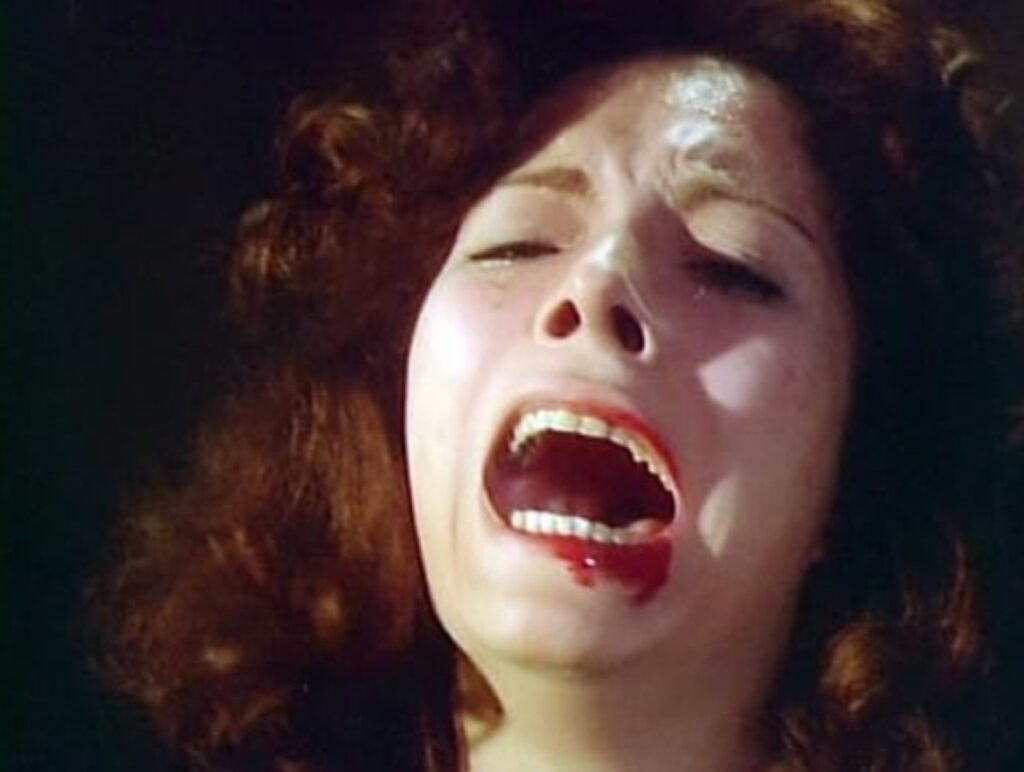 A close-up shot of Justine (Susana Kamini) screaming in agony and terror, her head tilted back with blood trickling down her chin and jaw against a dark background.