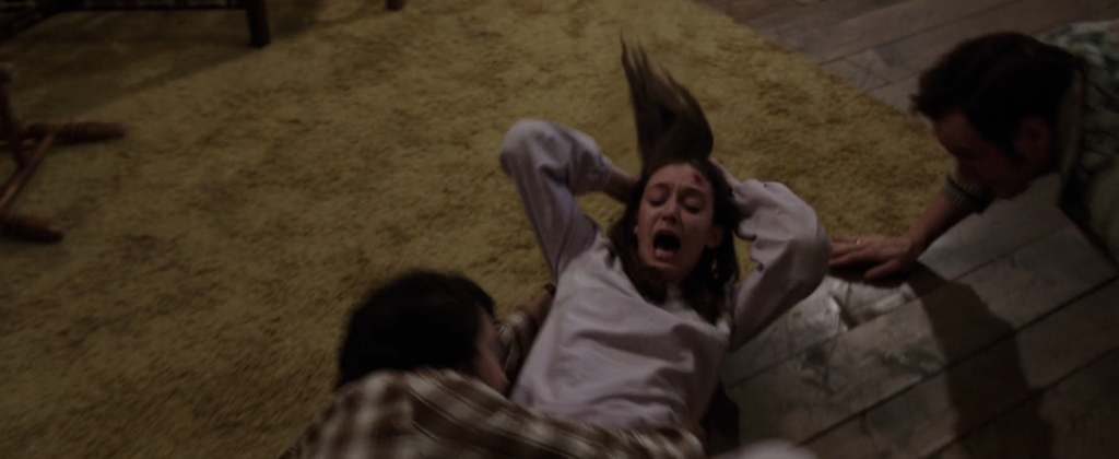 A teenage girl is on the floor. She is being pulled by her hair by an invisible force. Her hair is up but there is no hand on her hair. Two men are trying to hold her down so she can stop being dragged across the floor. She looks very scared.